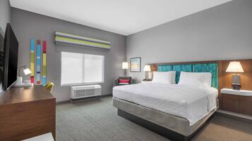 A hotel room with a large bed, a desk with a TV, a window with blinds, and a wall-mounted air conditioner.