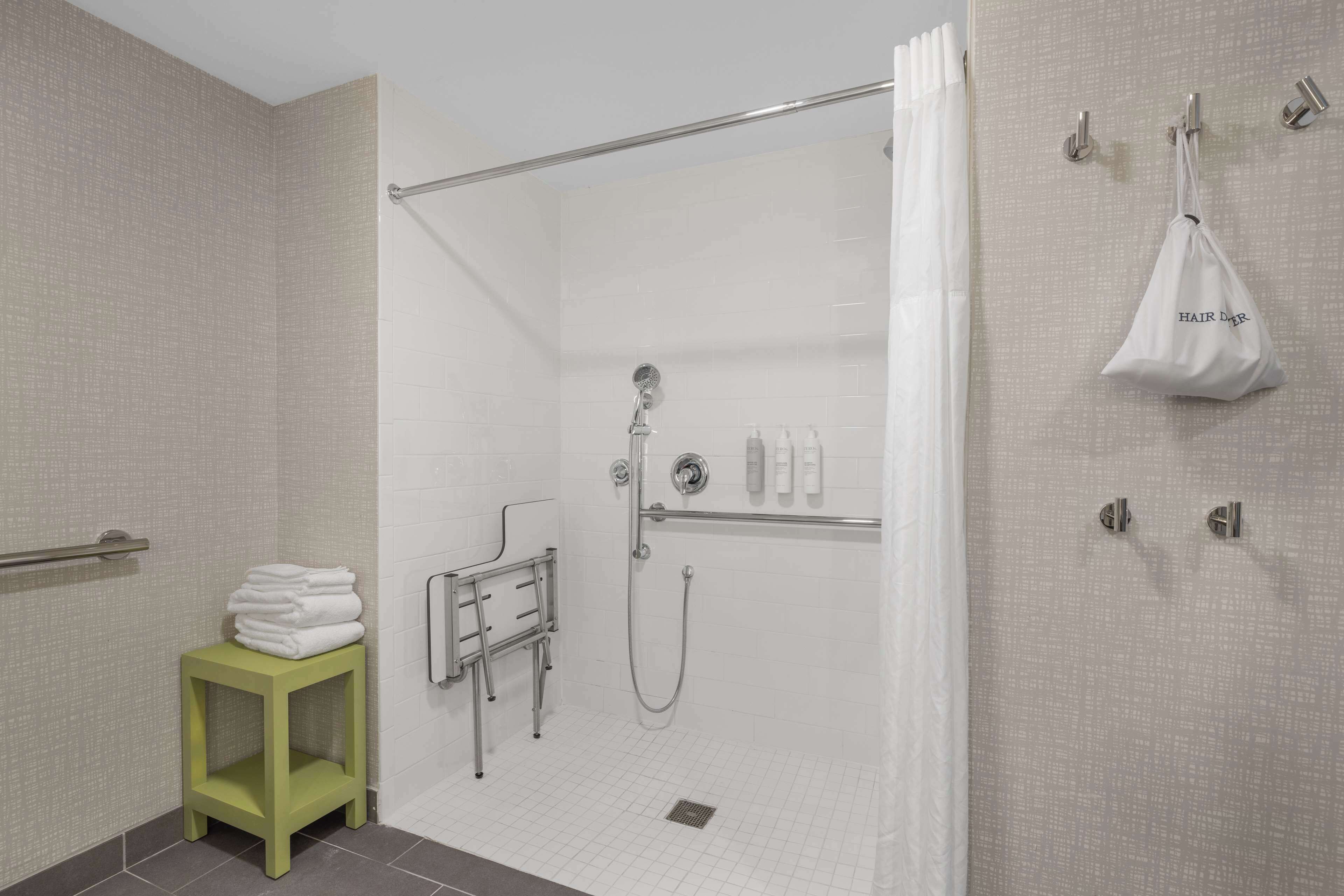 A modern bathroom with a shower, a green stool with towels, and a white bag hanging on hooks.