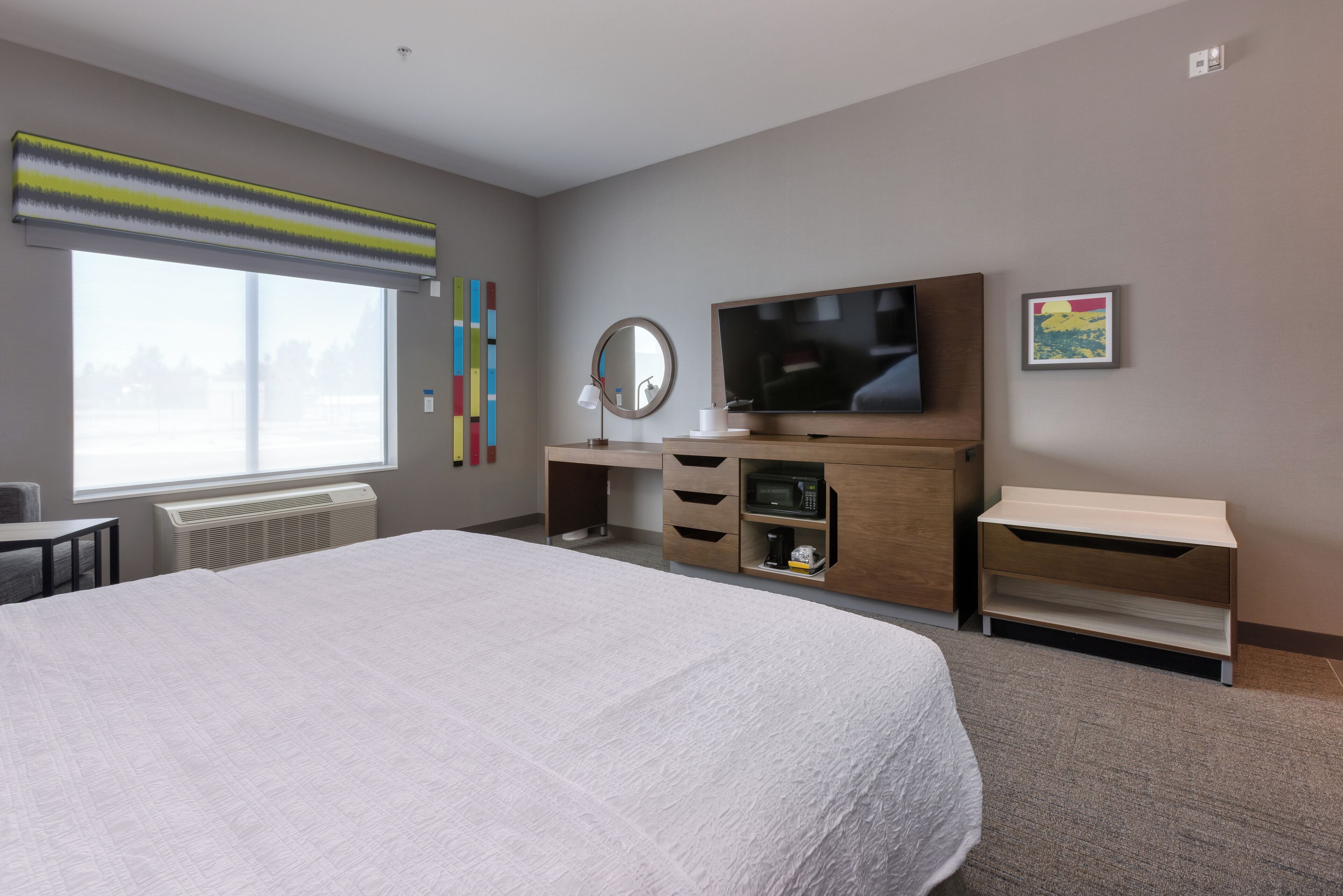 A hotel room with a large bed, a desk with a flat-screen TV, a mirror, and a small shelf with a microwave.