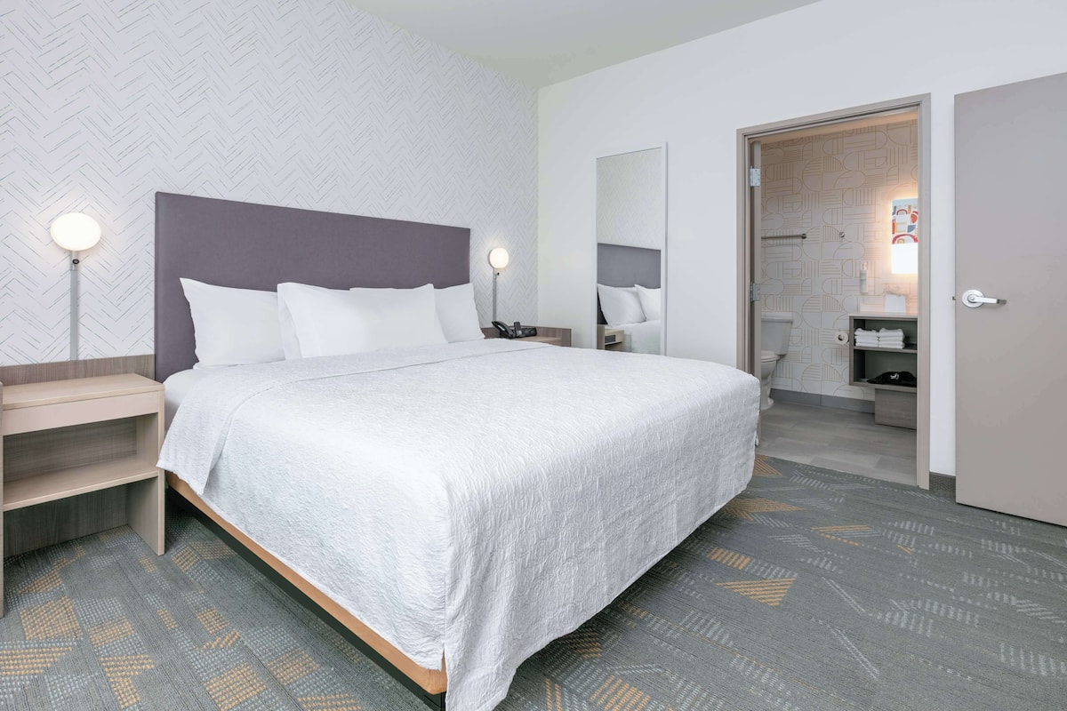 A modern hotel room with a large bed, bedside lamps, a wardrobe, and a bathroom visible through an open door.