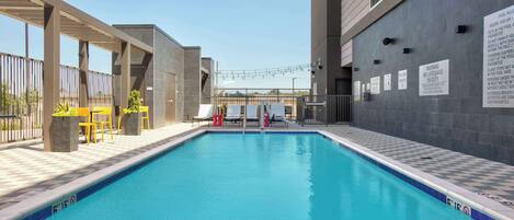 Une piscine dont le bord est orné d’un motif à carreaux, entourée d’une terrasse couverte aménagée avec des chaises et des tables, et un bâtiment à la façade grise sur lequel est affiché un panneau indiquant les règles d’utilisation de la piscine.