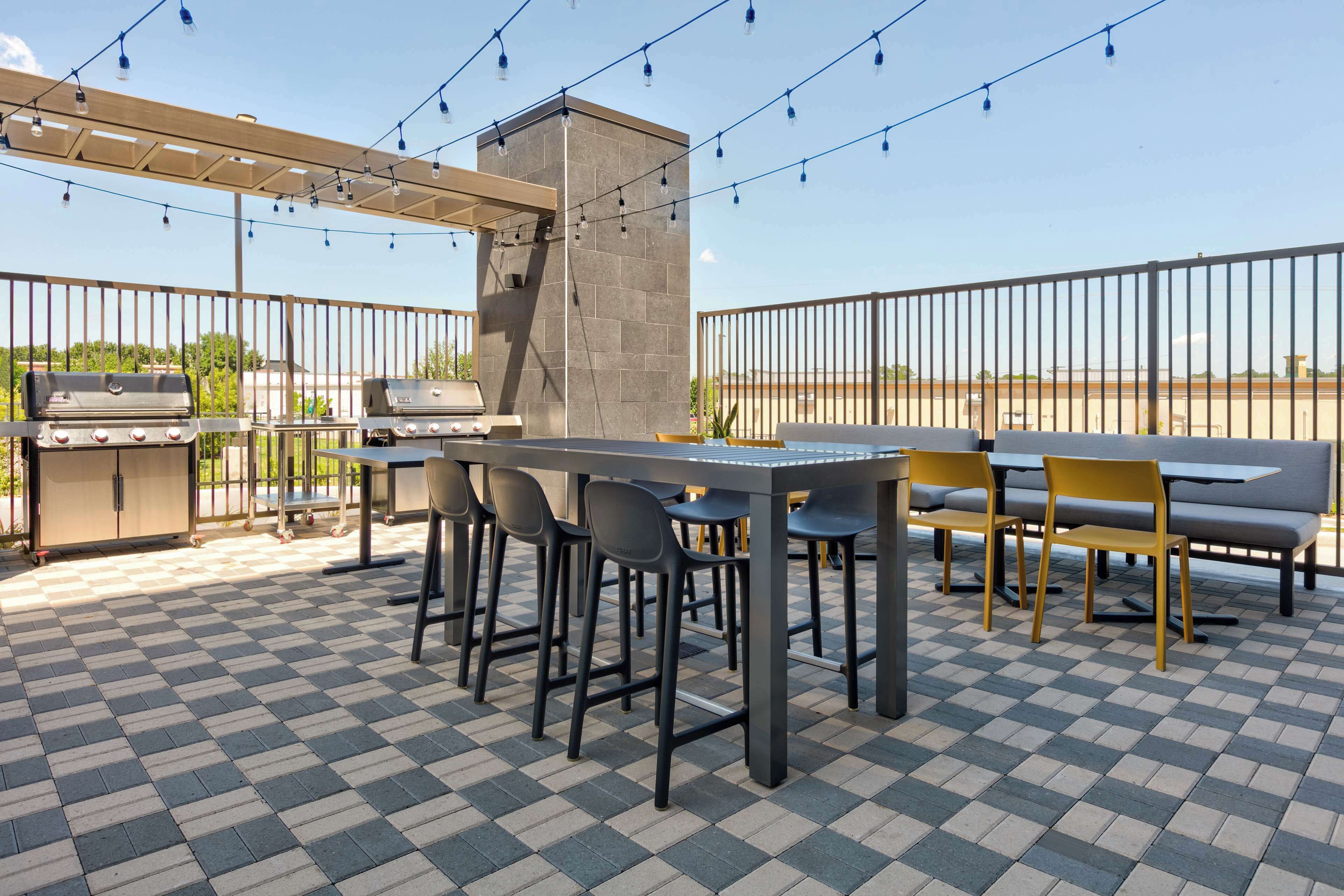 An outdoor patio with a checkered floor, a barbecue grill, a table with chairs, and a pergola with string lights.