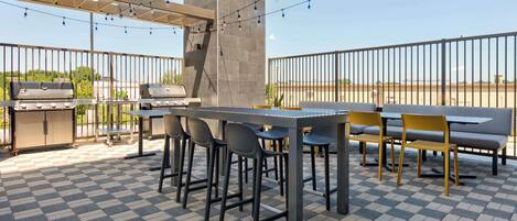 An outdoor patio with a checkered floor, a barbecue grill, a table with chairs, and a pergola with string lights.