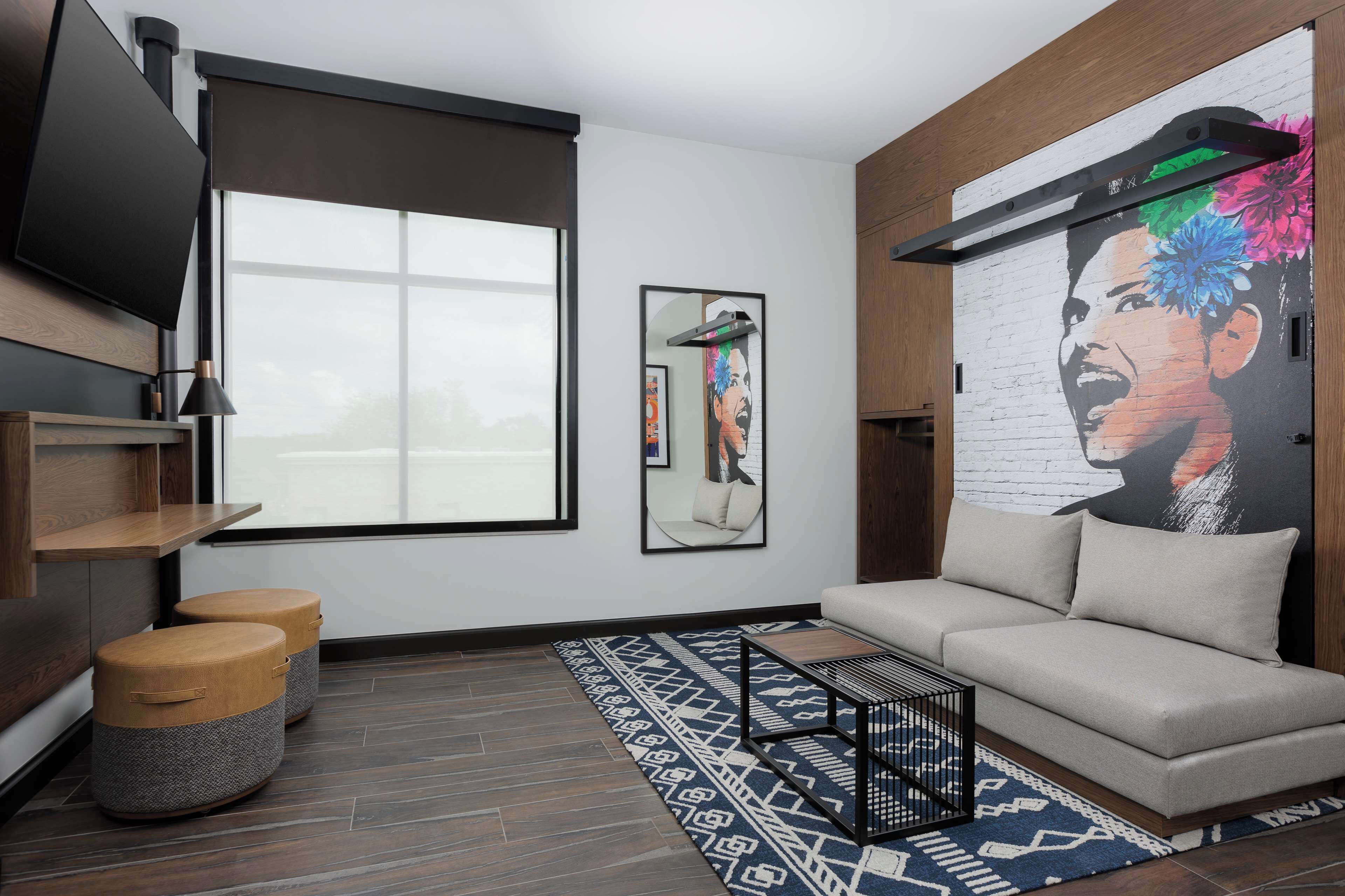 Una sala moderna con un sofá, una mesita de centro y dos otomanas. Hay un ventanal grande, un televisor montado en la pared y un mural en la pared.