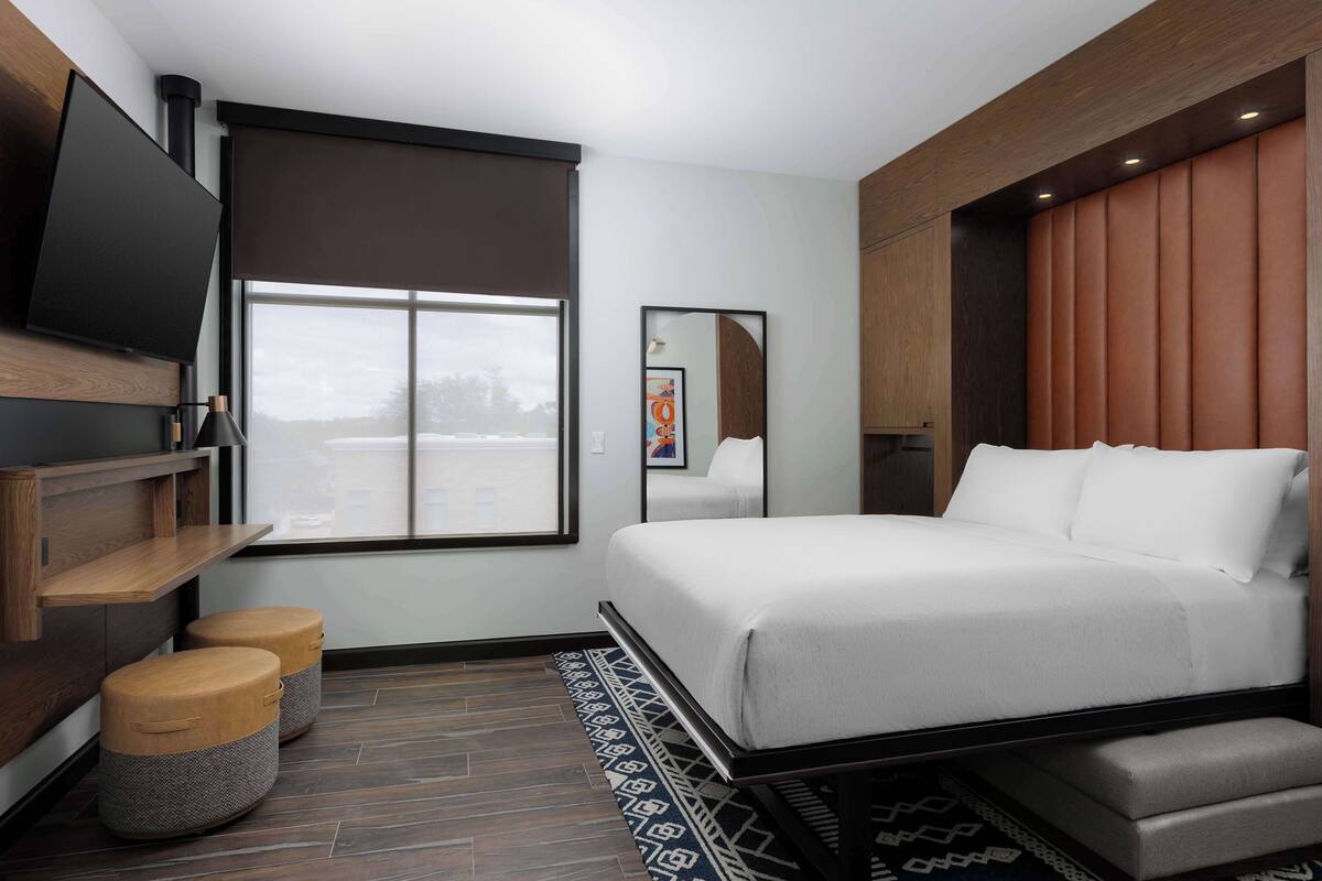 A modern hotel room with a large bed, a flat-screen TV mounted on the wall, a window with a view, and a wooden bench.