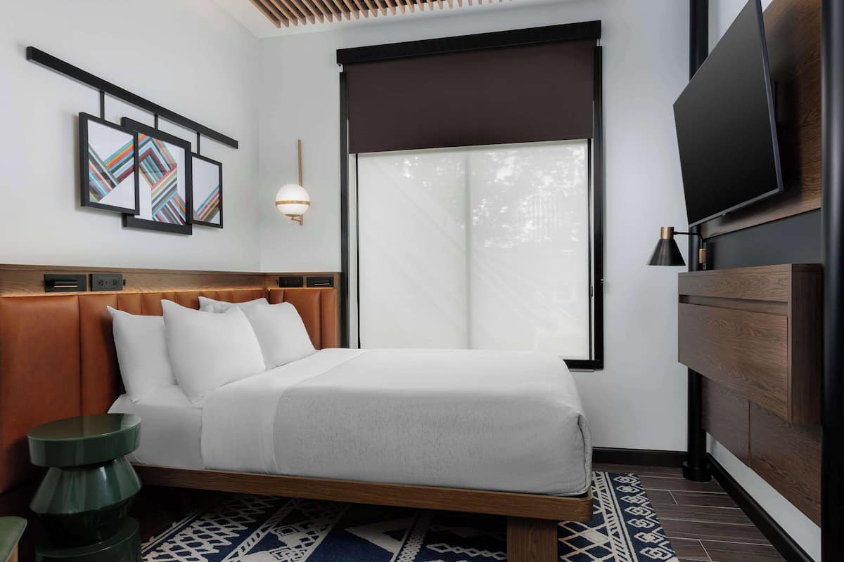 A modern hotel room with a bed, a television, a wall-mounted artwork, and a large window.