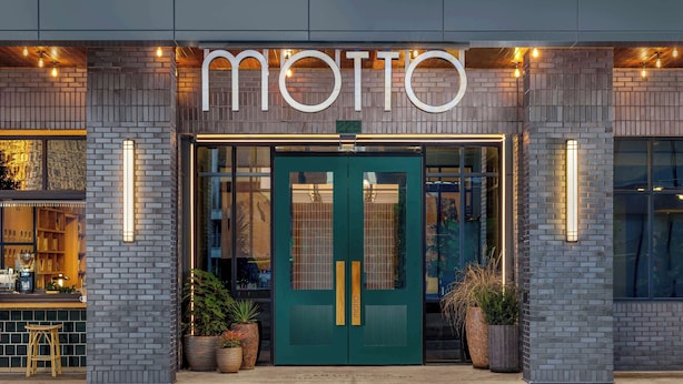 A building facade with a prominent "MOTTO" sign above a green door, flanked by potted plants and illuminated by wall-mounted lights.
