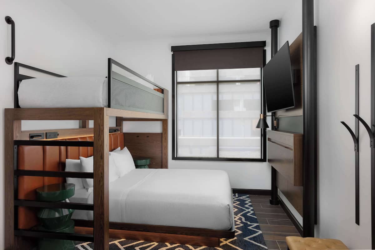 A compact hotel room with a bunk bed, a single bed, a TV, and a window with blinds.
