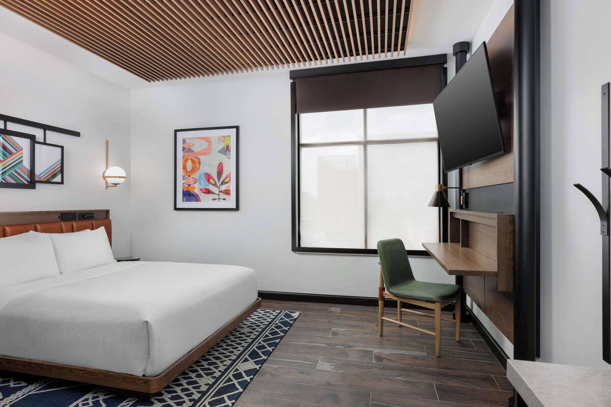 A hotel room with a large bed, a desk, a chair, a TV, and artwork on the walls.