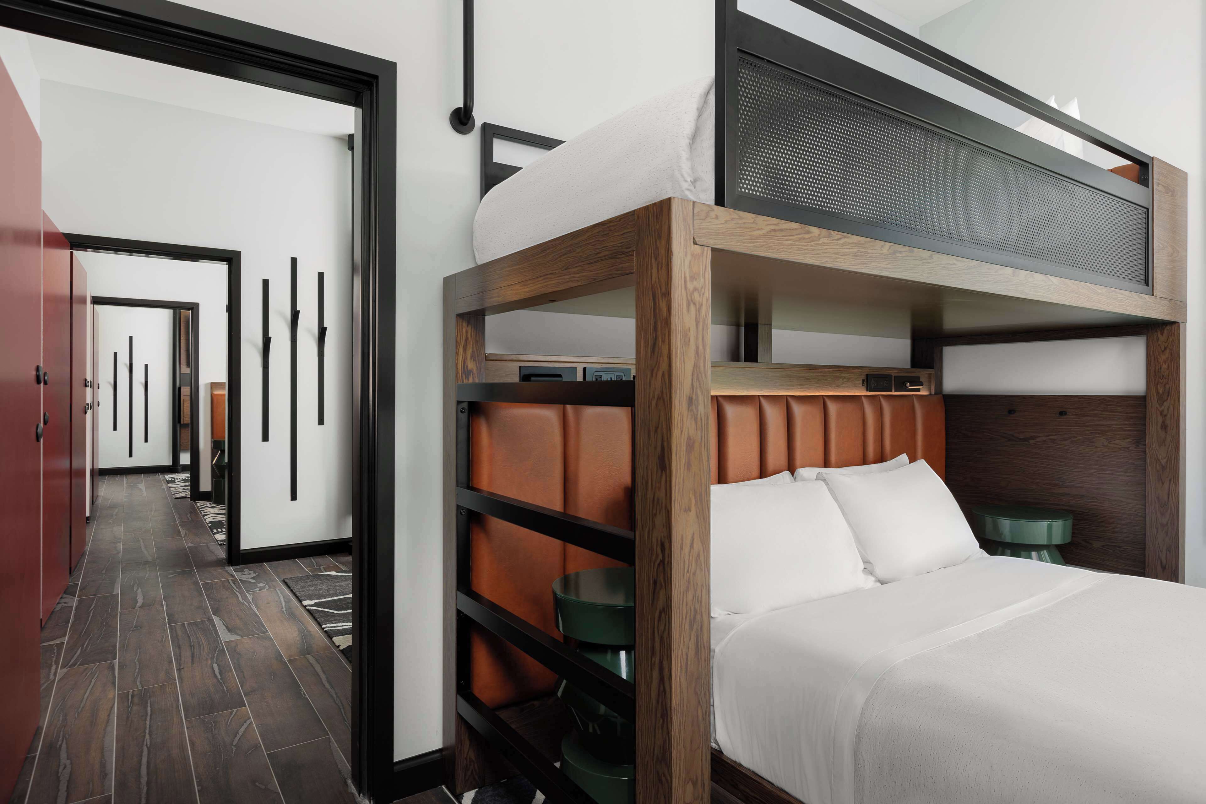 A modern hotel room with a bunk bed, a wooden headboard, a small green side table, and a large window.