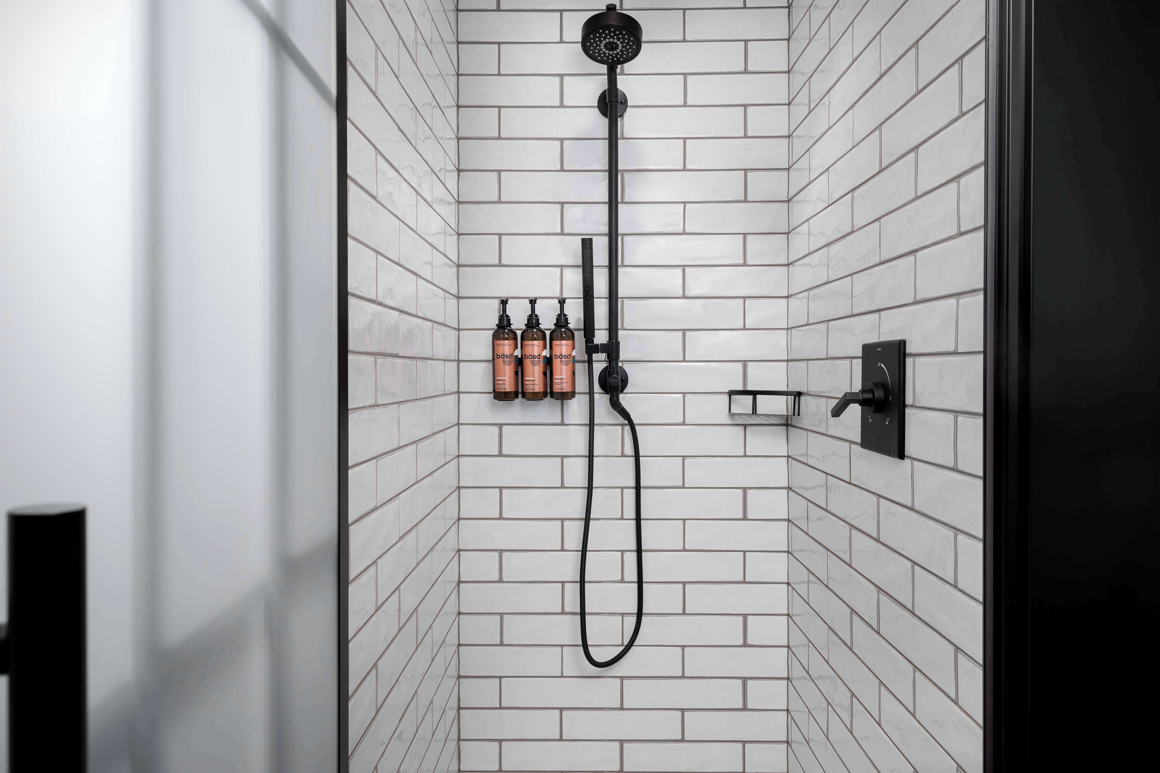 A modern shower with a black hose and wall-mounted controls, white tiled walls, and three bottles of shampoo and conditioner.
