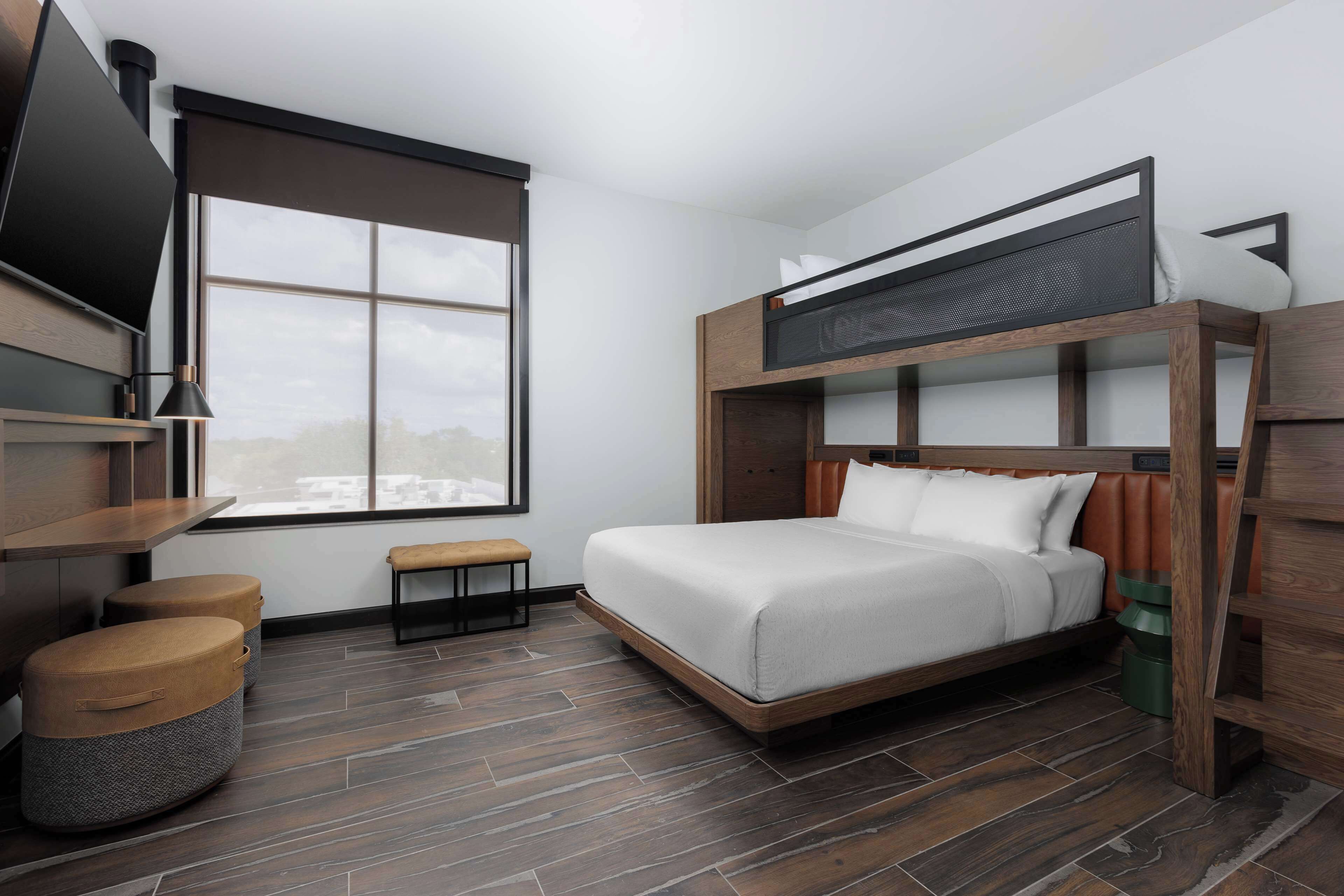 Una habitación de hotel moderna con literas, un televisor de pantalla plana montado en la pared, un banco y una ventana con vista a la ciudad.