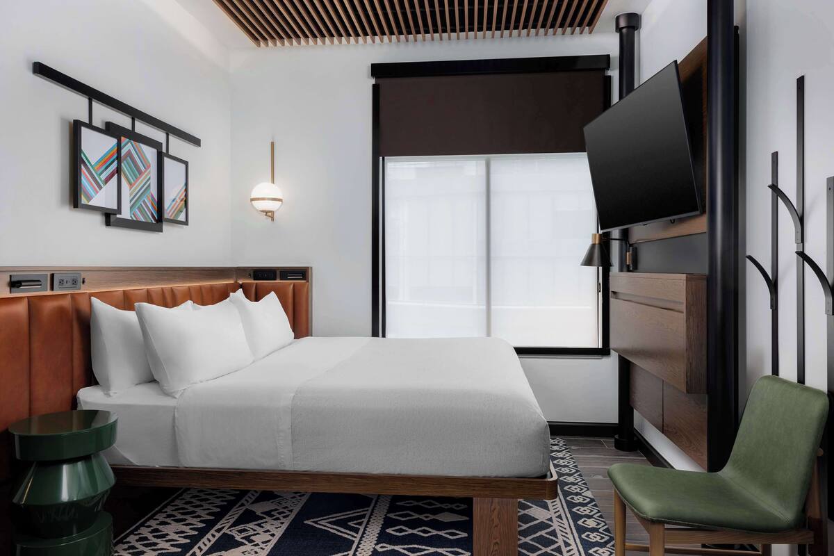 A hotel room with a bed, a green chair, a TV, a wall-mounted artwork, and a window with blinds.