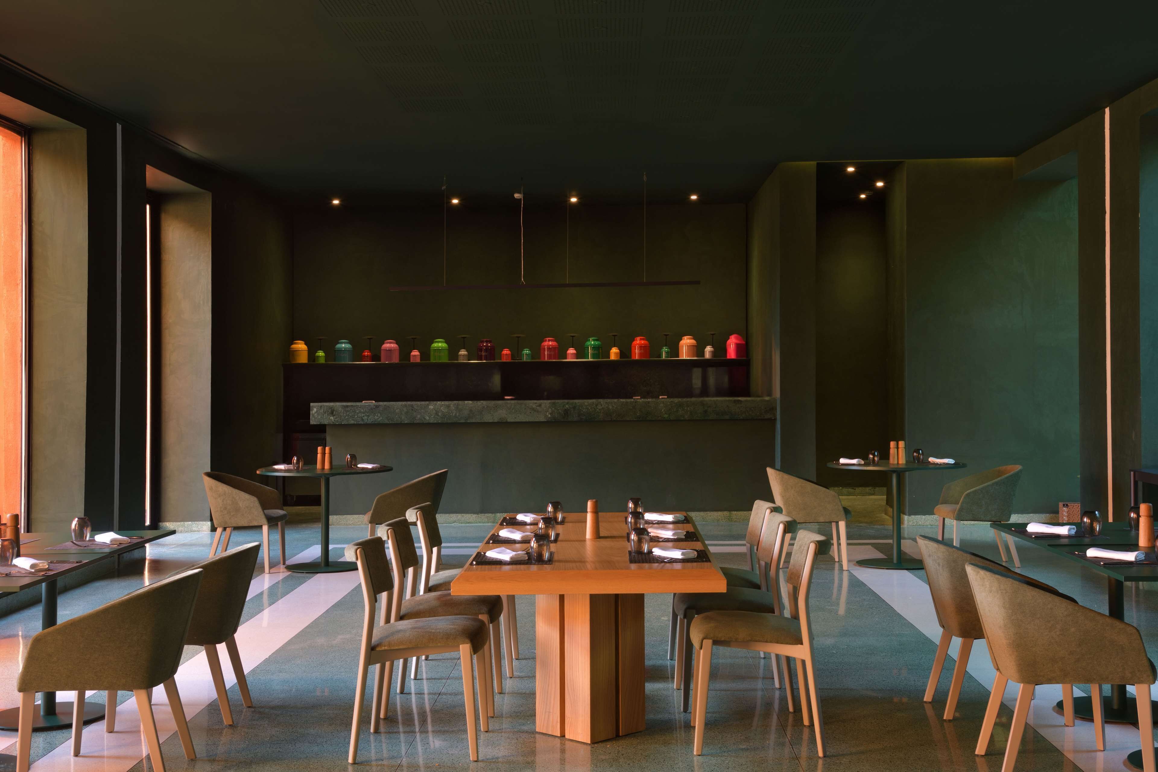 A modern dining area with a central wooden table surrounded by chairs, a long shelf with colorful bottles, and a minimalist design.