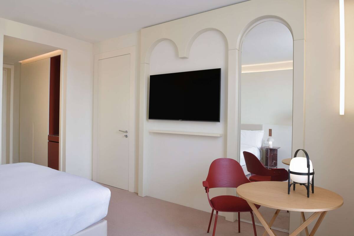 Une chambre d’hôtel avec un lit, un téléviseur fixé au mur, une chaise rouge et une petite table ronde.