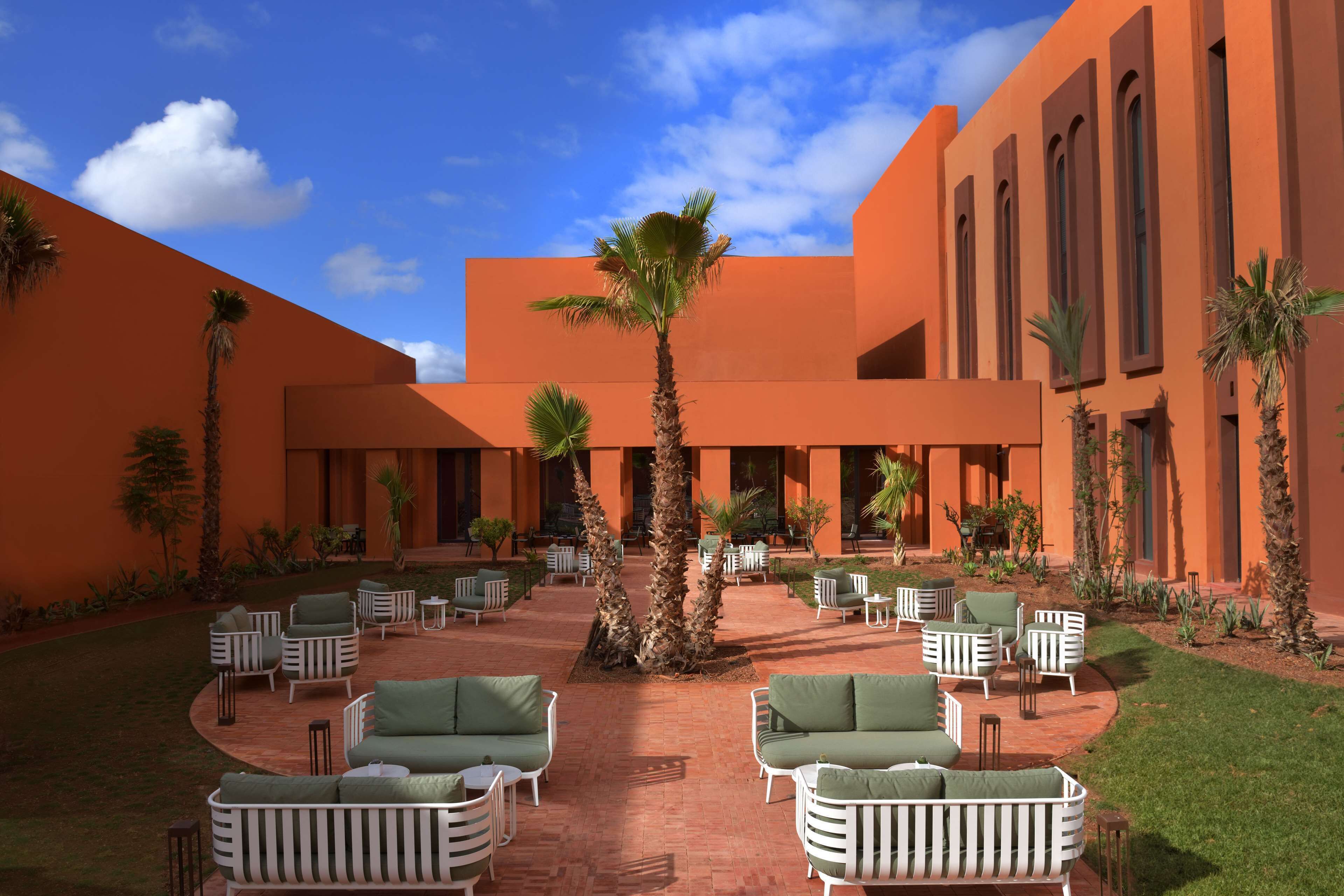 Un patio con mesas al aire libre, palmeras y un edificio de paredes anaranjadas.