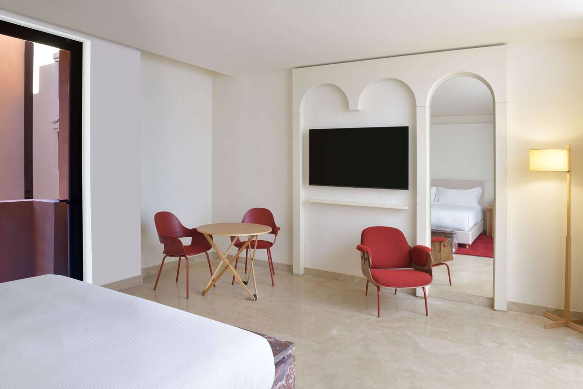 Une chambre d’hôtel avec un lit, un téléviseur fixé au mur, une petite table ronde avec deux chaises rouges et un miroir surmonté d’un arc.