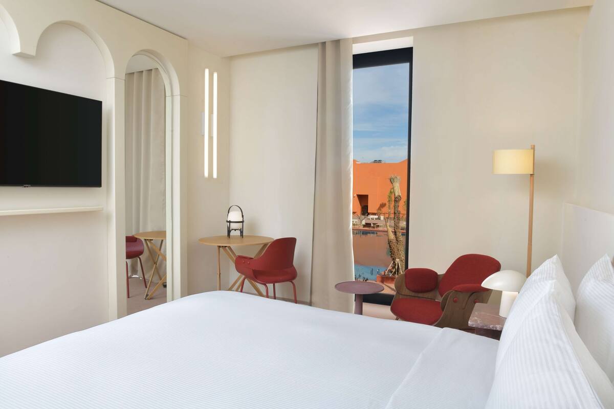 Une chambre d’hôtel avec un grand lit, un téléviseur fixé au mur, une petite table avec des chaises et une vue sur l’extérieur.