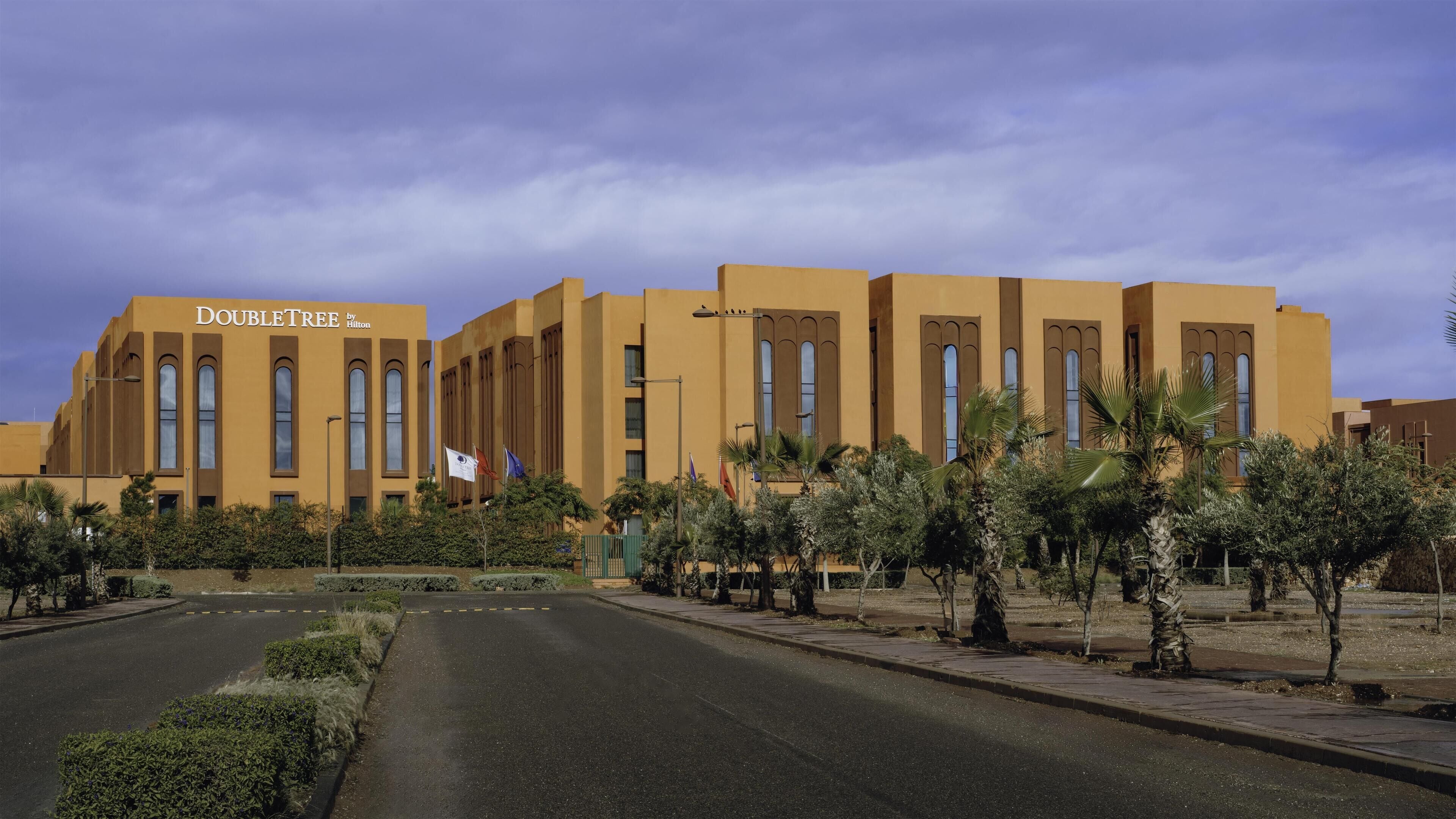 Un hotel llamado "DOUBLETREE by Hilton" con un diseño arquitectónico moderno, destacando amplios ventanales y un jardín cuidado con palmeras.