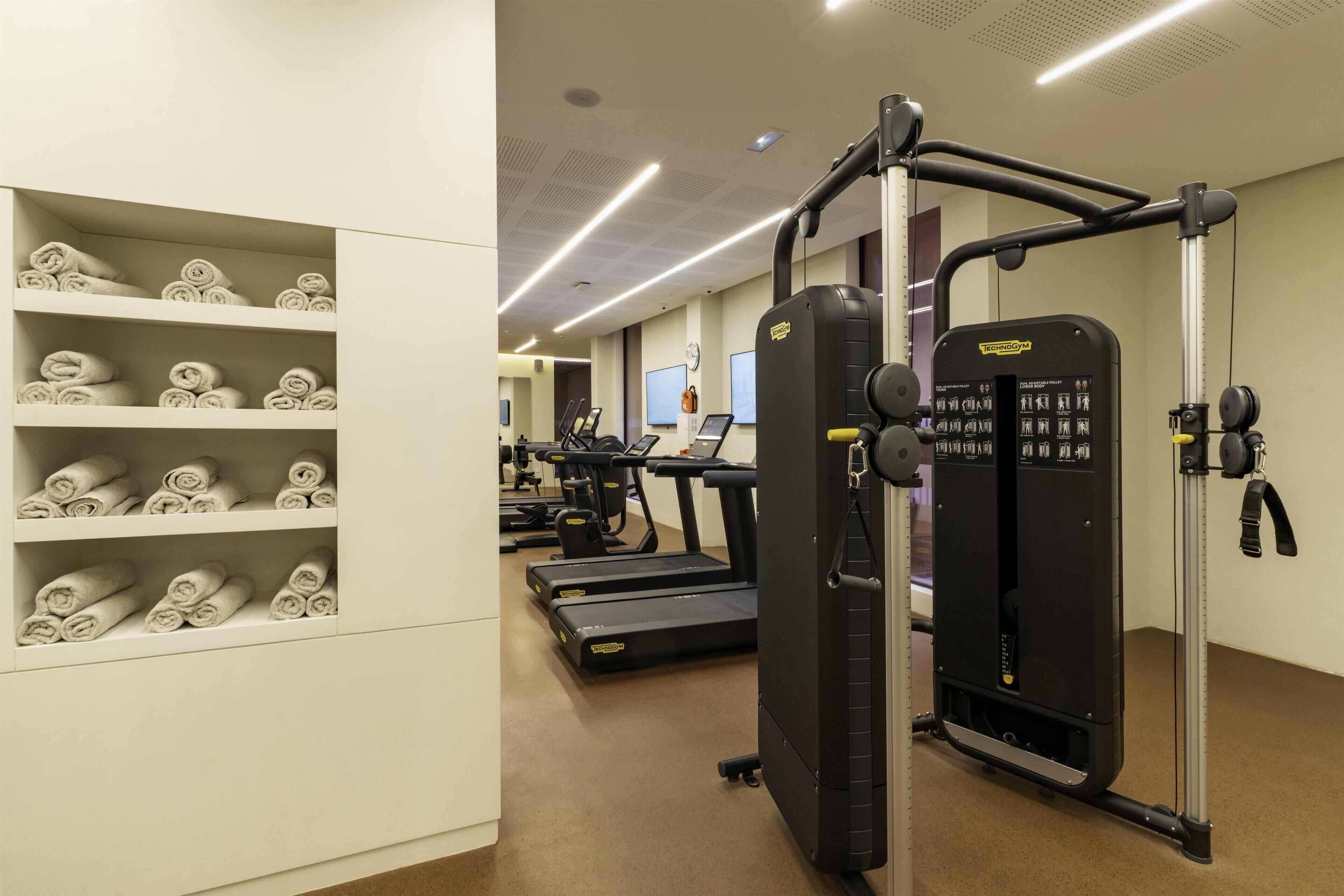 A gym with exercise equipment, including treadmills and a weight machine, and shelves with rolled towels.