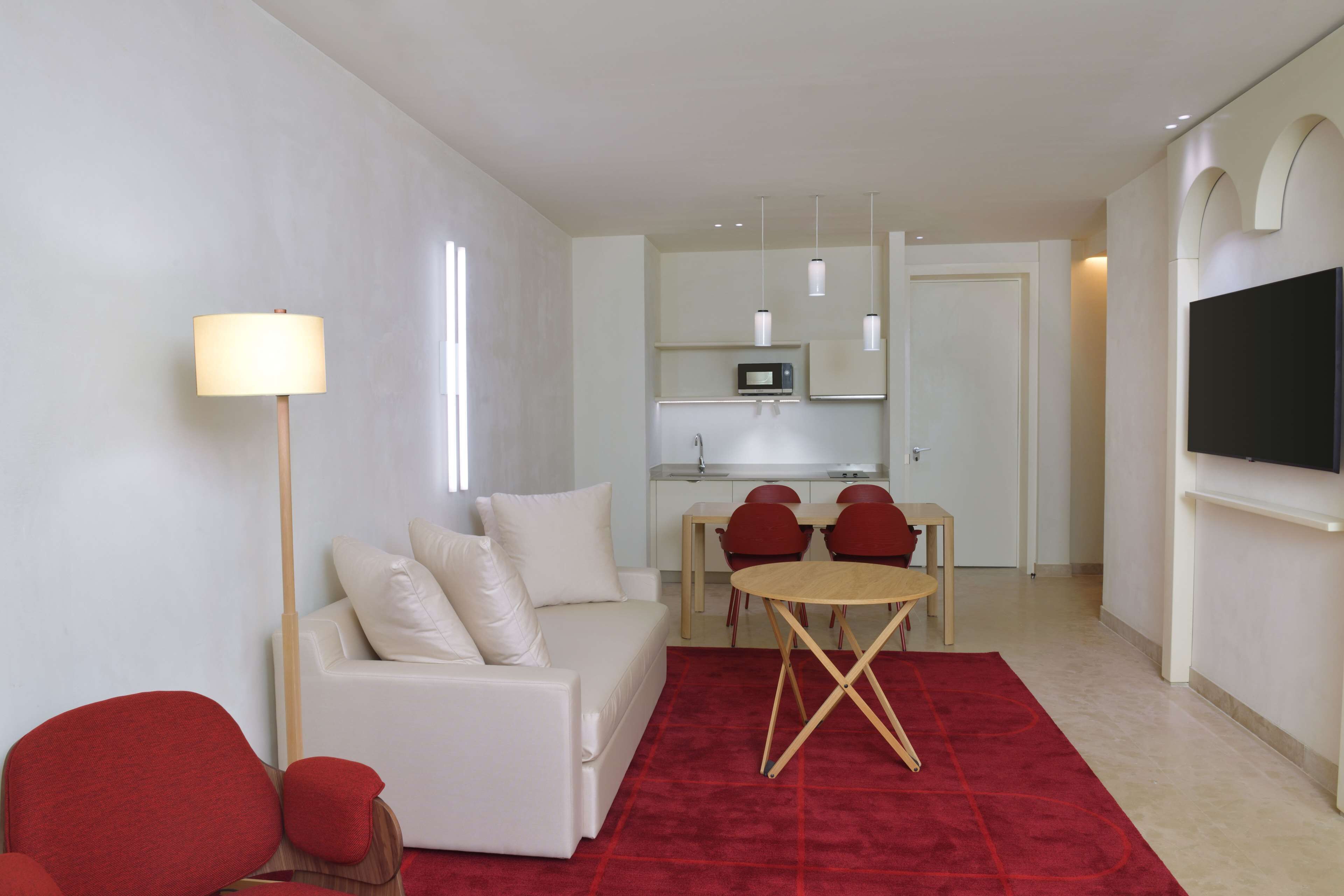 A modern living room with a white sofa, red chairs, a red carpet, a dining table, a TV, and a kitchen area with a microwave.