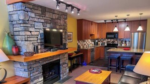 Una sala moderna con chimenea de piedra, televisor de pantalla plana, muebles de madera y una zona de cocina con electrodomésticos de acero inoxidable.