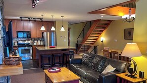 Una cocina moderna con electrodomésticos de acero inoxidable, un comedor con una mesa y sillas de madera, un sofá de cuero y una escalera que conduce a un nivel superior.