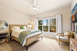 Un dormitorio con cama, escritorio, silla, ventilador de techo y una ventana grande con vistas a árboles.
