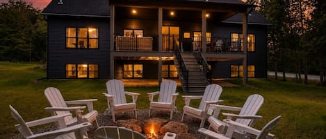 A two-story house with a fire pit and Adirondack chairs in the front yard.