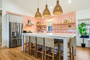 A modern kitchen with a stainless steel refrigerator, a central island with bar stools, wicker pendant lights, and pink-tiled backsplash.