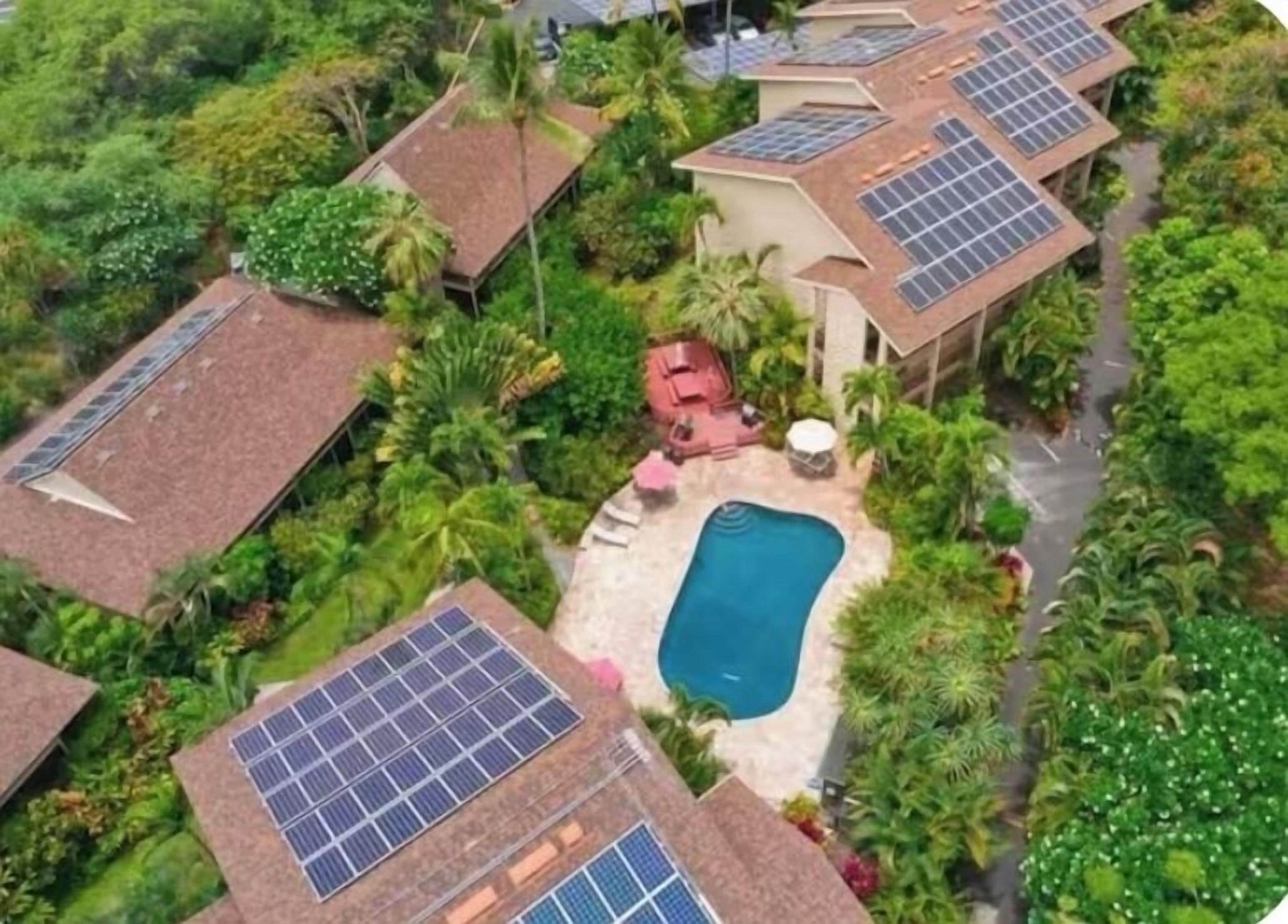 A residential area with multiple houses, each equipped with solar panels on the roofs. There is a swimming pool in the center surrounded by greenery.