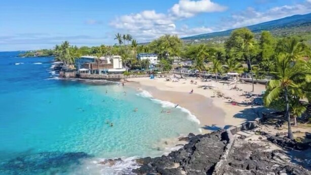 A coastal beach with clear turquoise water, a sandy shore, and a few buildings including a prominent white structure with a blue roof. - Peaceful Retreat • 5 min Walk to Town, Pier & Beach • Free Parking (Kailua-Kona)
