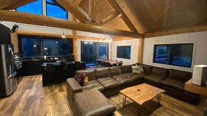 A spacious living area with a wooden ceiling, a large leather sofa, a wooden coffee table, and a kitchen area with a refrigerator and stove. - Timberwolf A Cabin at Sasquatch Mountain  *HOT TUB* GAMES ROOM* (Agassiz)