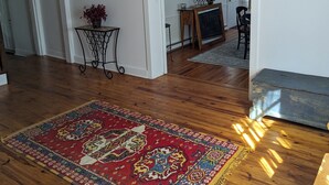 Un salon spacieux doté d’un parquet, d’un grand tapis rouge à motifs, d’un coin repas avec une table et des chaises, et d’une horloge au mur.