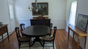 Une salle à manger avec une table ronde, quatre chaises, un lustre, un tableau au-dessus de la table et un banc en bois avec un cadre photo sur le côté.