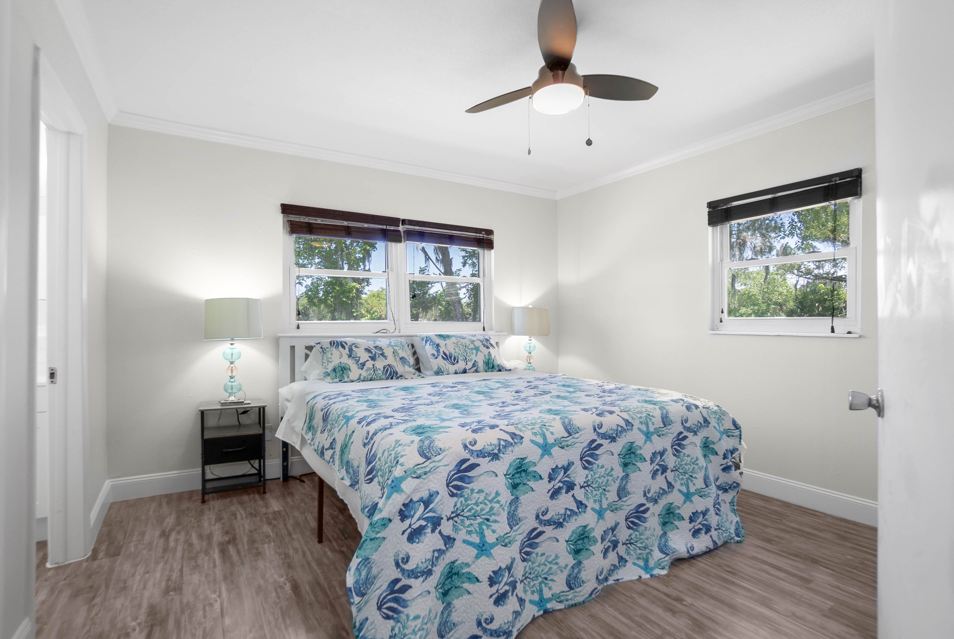 A bedroom with a bed, two bedside lamps, a ceiling fan, and two windows with blinds.