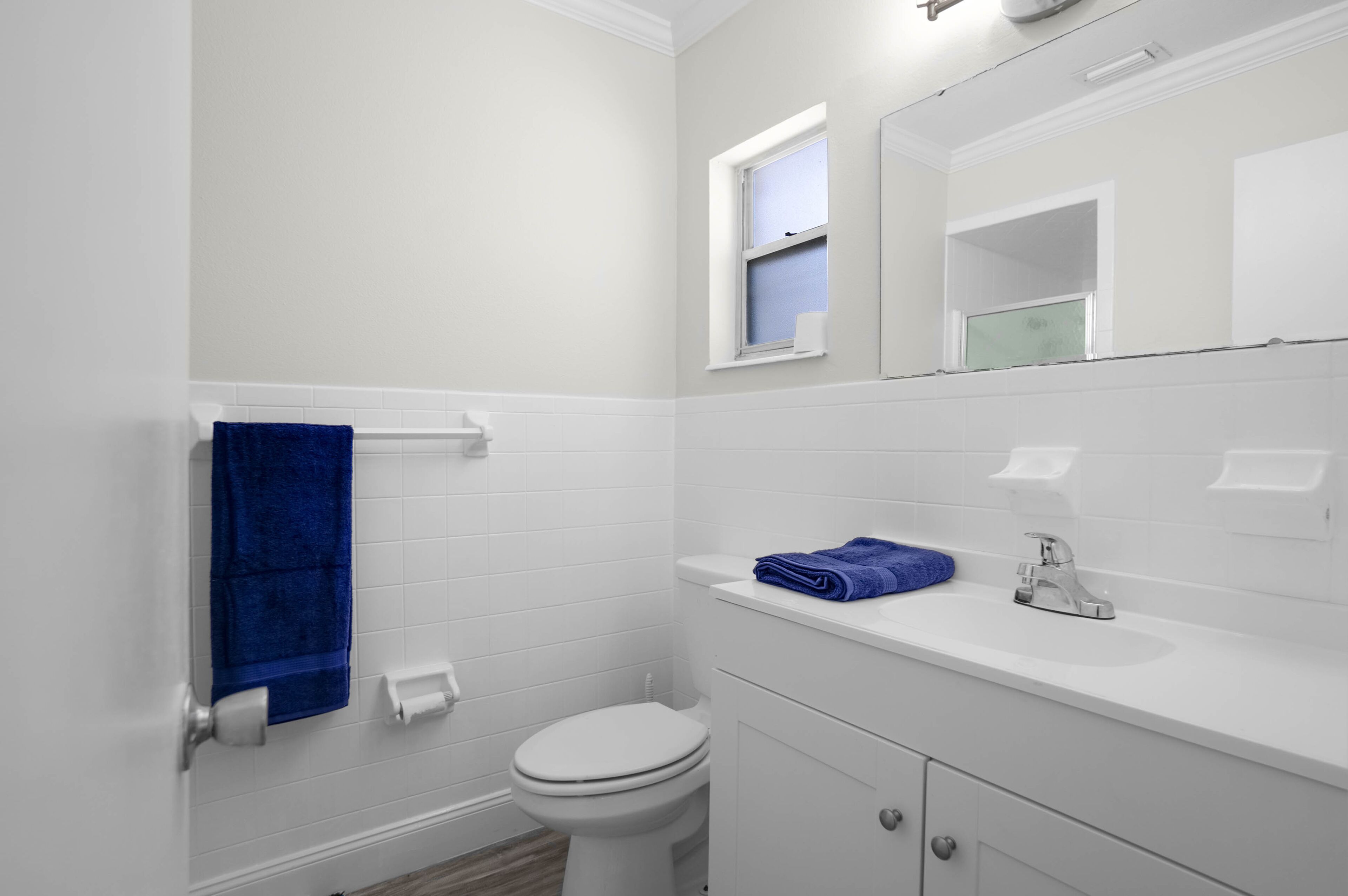Casa de banho com azulejos brancos, sanita, lavatório com espelho e toalha azul pendurada na parede.