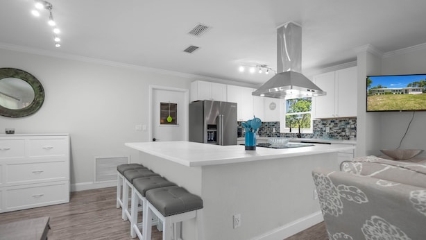 A modern kitchen with white cabinetry, a central island with bar stools, a stainless steel refrigerator, and a large window overlooking a garden. - Welcome to the scallop shack! Newly renovated and on the water in Crystal River! (Crystal River)