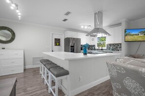 Una cocina moderna con armarios blancos, una isla central con taburetes y un refrigerador de acero inoxidable, y un amplio ventanal con vistas a un jardín.