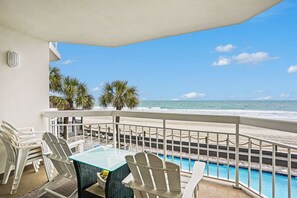 A balcony with white chairs and a glass table overlooking a beach with palm trees and a pool.