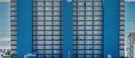 A large hotel with a central tower, multiple floors, and a parking lot with several cars.