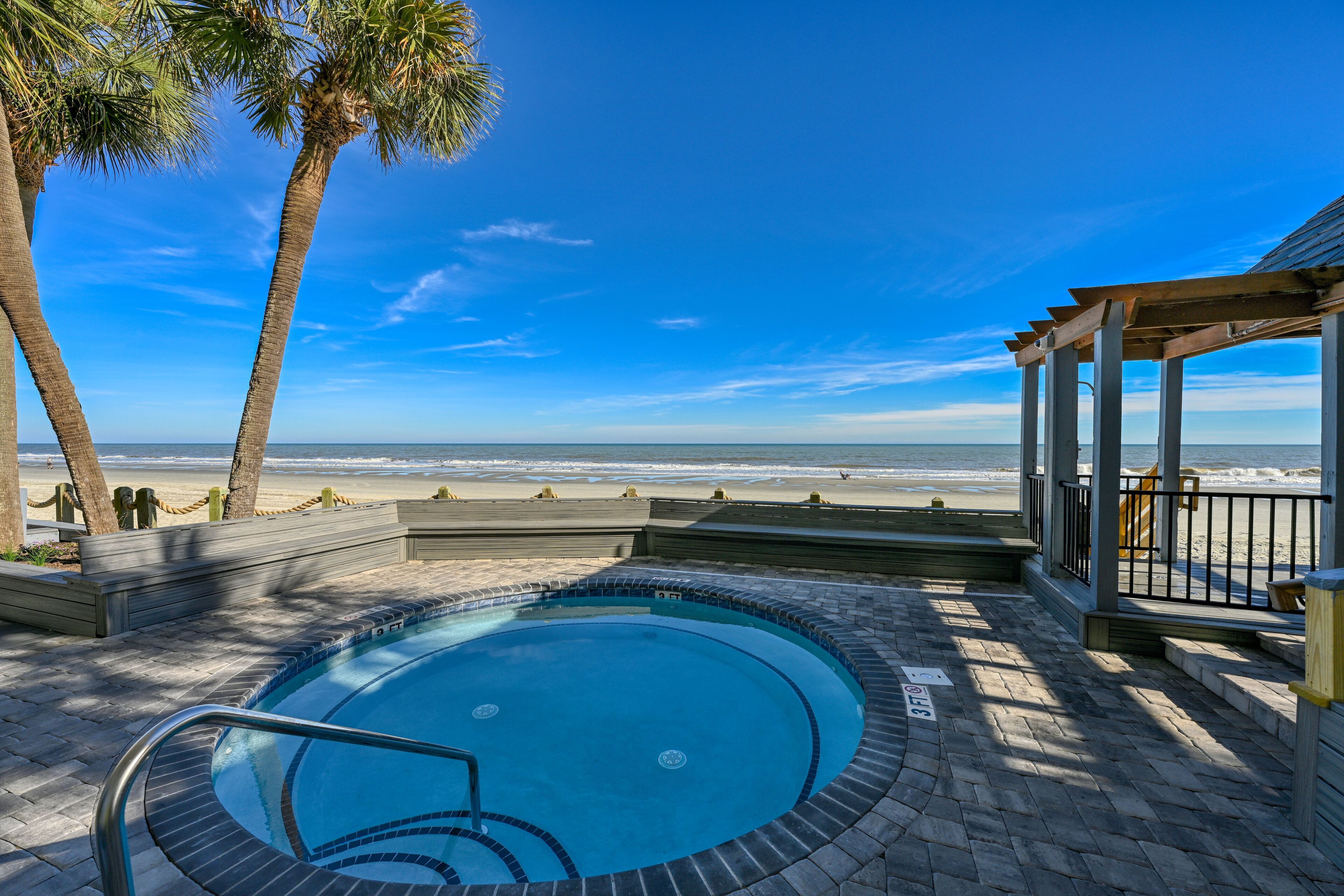 A pool area with a view of the beach, palm trees, and a covered seating area.