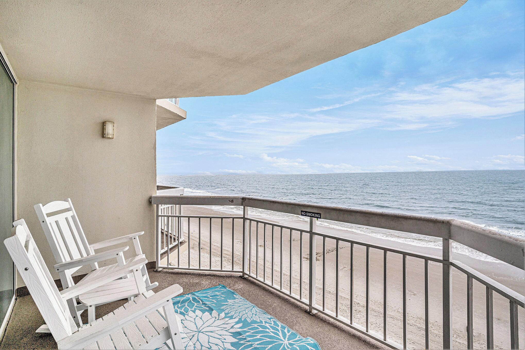 A balcony with a view of the ocean, white chairs, and a blue patterned rug.