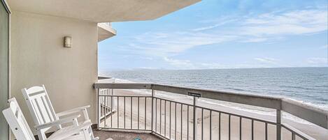 A balcony with a view of the ocean, white chairs, and a blue patterned rug.