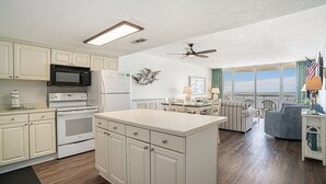 A kitchen with white cabinets, a microwave, and an oven. There is a dining area with a table and chairs, and a view of the ocean through large windows. - Oceanfront 2bed/2bath-Water's Edge Resort-711 Indoor/Outdoor Pool & Hot Tub (Garden City Beach)