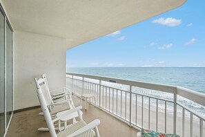 A balcony with white wicker chairs and a small table, overlooking a beach with a 'NO PARKING' sign.