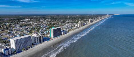 A coastal cityscape with a beachfront hotel featuring multiple floors, a sandy beach, and a clear blue sky.