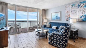 A room with a balcony, a blue sofa, a patterned armchair, a small table, and a wall with nautical decor. - Oceanfront 1bed/2bath-Water's Edge Resort-1206 Indoor/Outdoor Pool & Hot Tub (Garden City Beach)