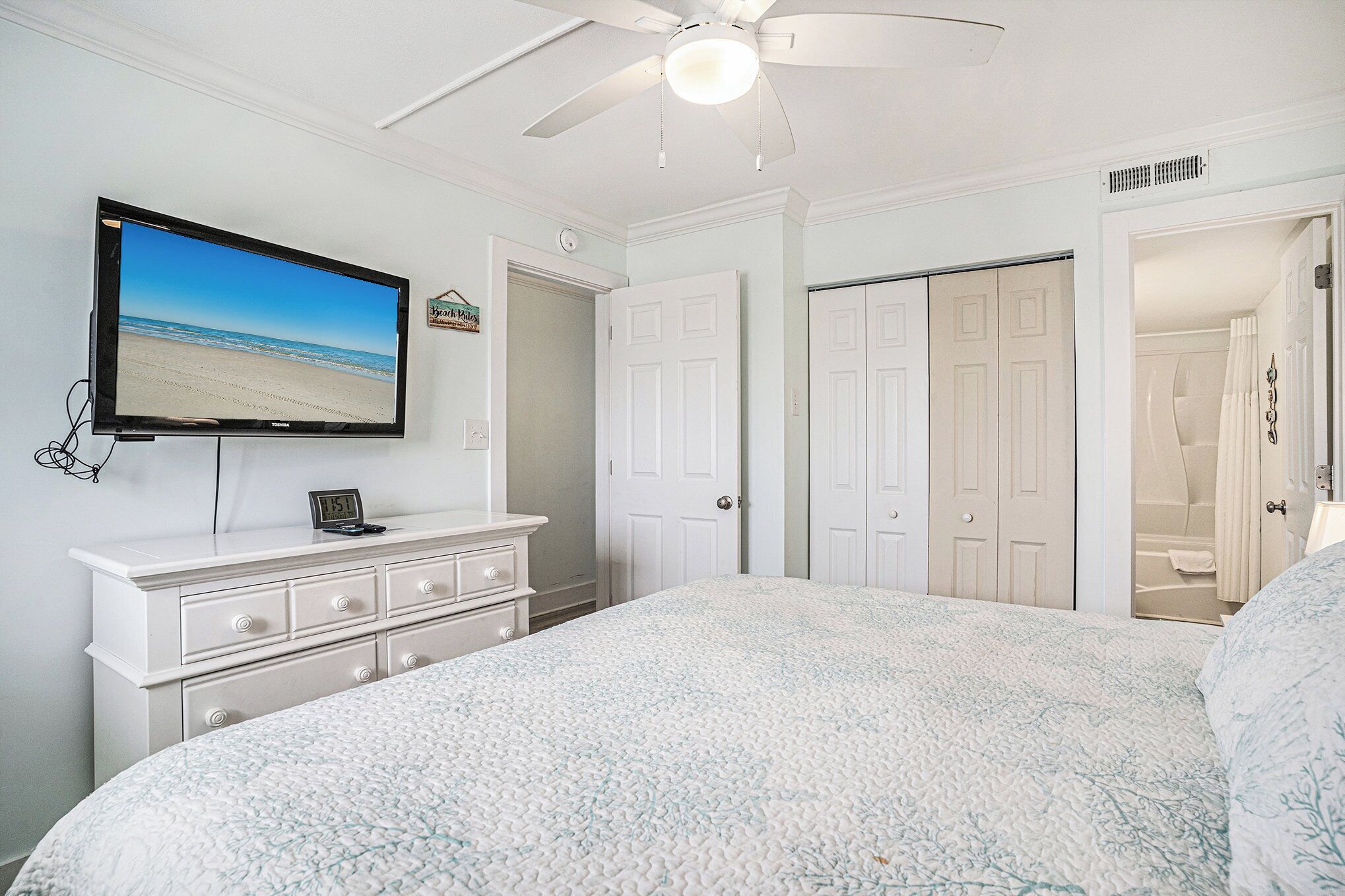 A bedroom with a bed, a TV on a dresser, a ceiling fan, and white doors.