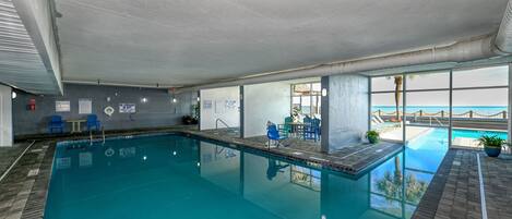 An indoor swimming pool with clear blue water, a view of the ocean through large windows, and a seating area with blue chairs.